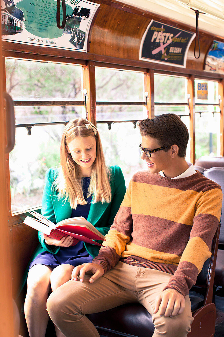 A young couple sitting on a bus looking at a book, her wearing a skirt and jacket and him wearing a jumper and trousers