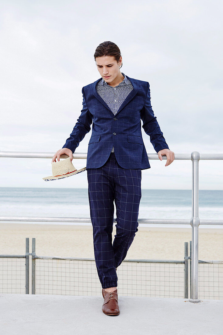 A young man on the beach wearing a printed shirt, a dark jacket and trousers