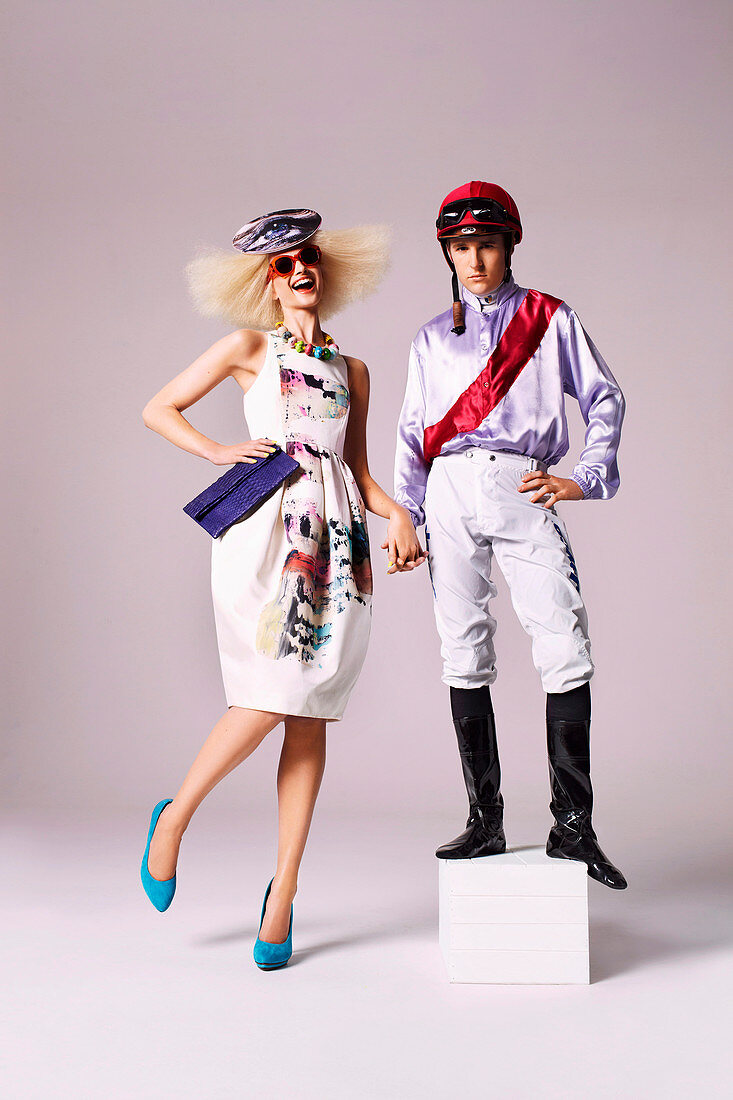 A blonde woman wearing a summer dress and a hat with a man in a jockey's outfit standing on a stool