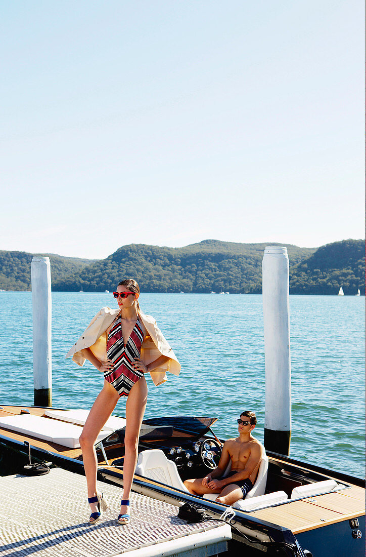 Junge Frau in gestreiftem Badeanzug und Blazer und junger Mann in Shorts auf einem Boot
