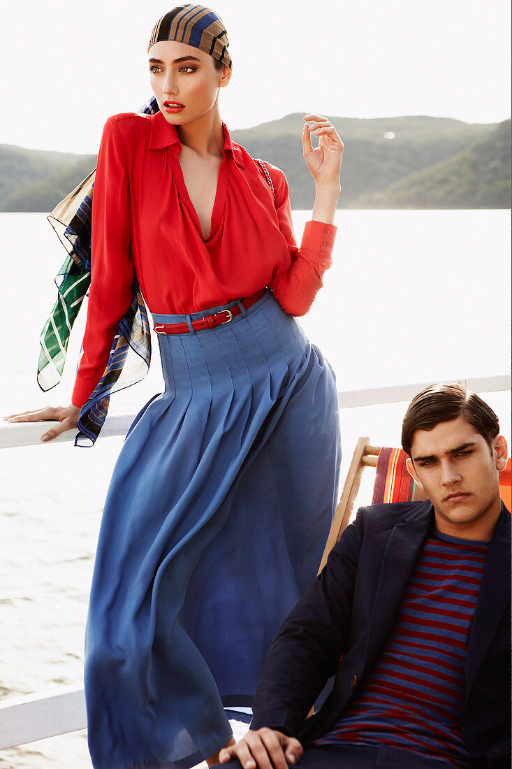 A young woman wearing a headscarf, a red blouse and a blue skirt standing next to a man sitting on a deckchair