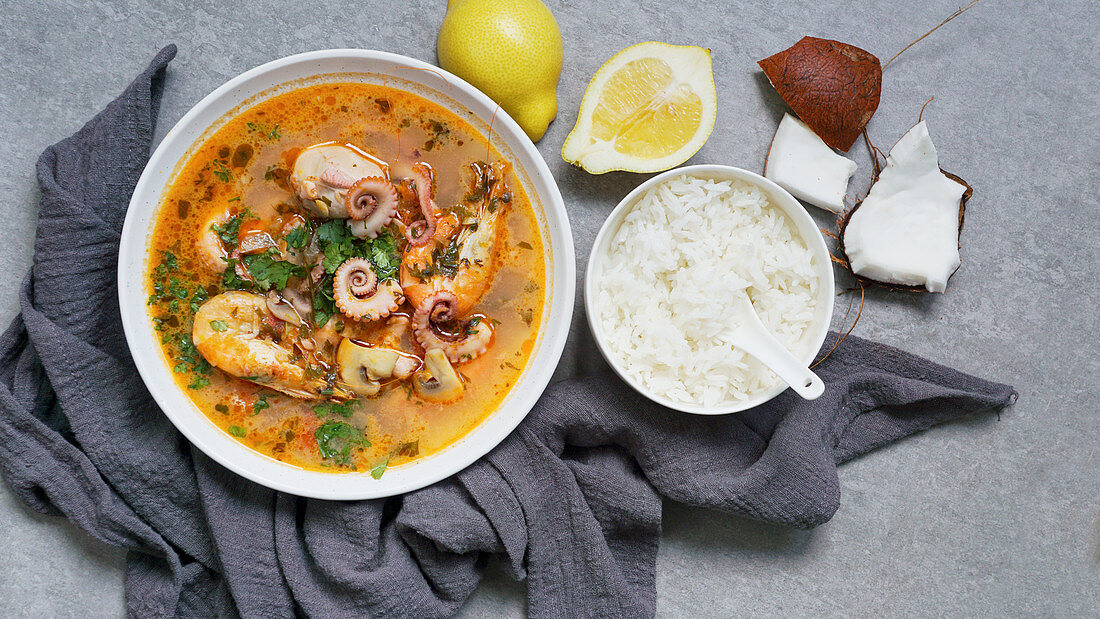 From above bowl of tom yum with shrimps, octopus and herbs near rice, lemon and coconut on grey background