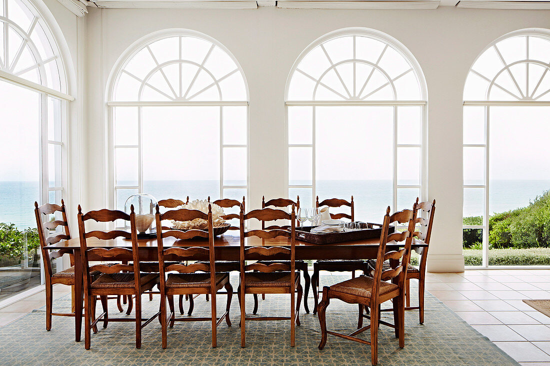 Lange Tafel mit Holzstühlen vor Rundbogen-Fensterfronten mit Meerblick