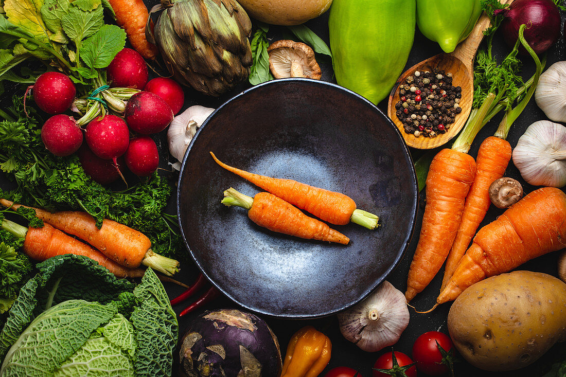 Two fresh farm carrots in vintage bowl and assorted organic vegetables on rustic black concrete background