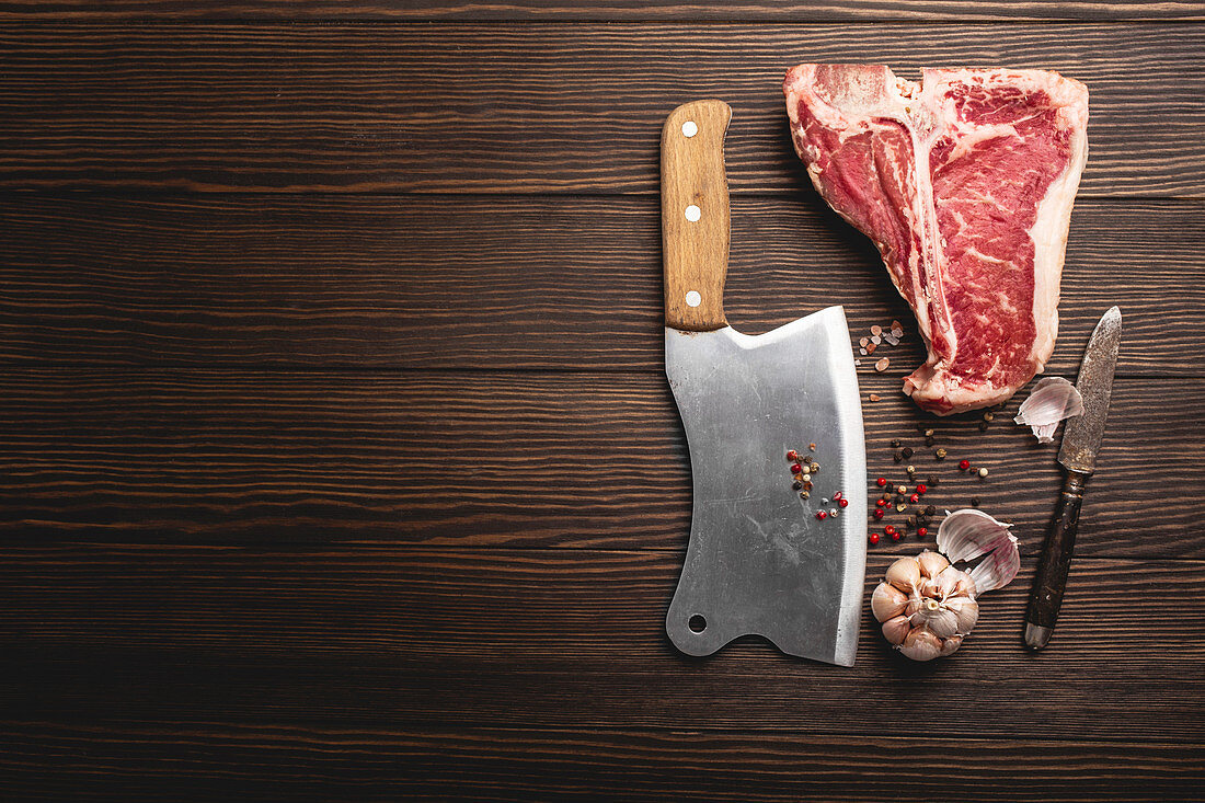 A raw T-bone steak with spices, a knife and a cleaver on a wooden surface