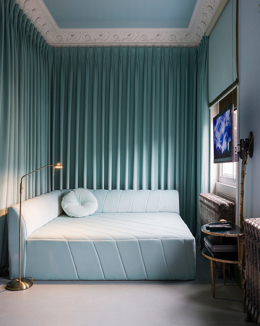 Square, pale blue sofa bed in corner with curtains, flatscreen TV and roller blind