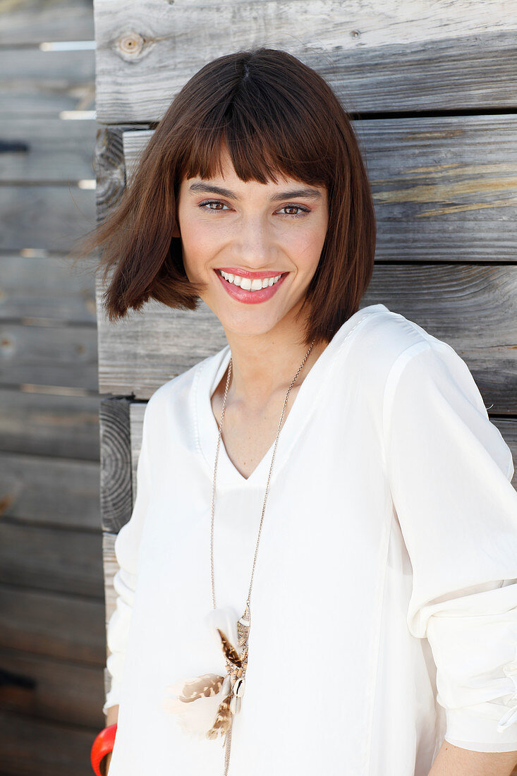 A brunette woman wearing a white long-sleeved blouse standing in front of a wooden wall