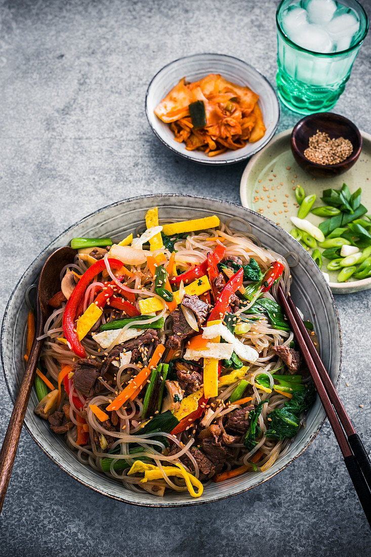 A bowl of korean glass noodles stir-fried with vegetables, topped with toasted sesame seed and garnished with spring onions and kimchi