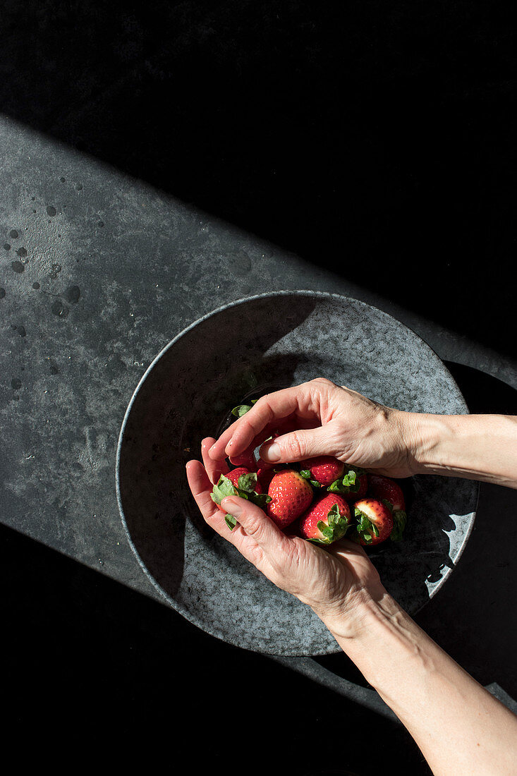 Hands holding pile of fresh strawberry near bowl illuminated by sunbeams in darkness
