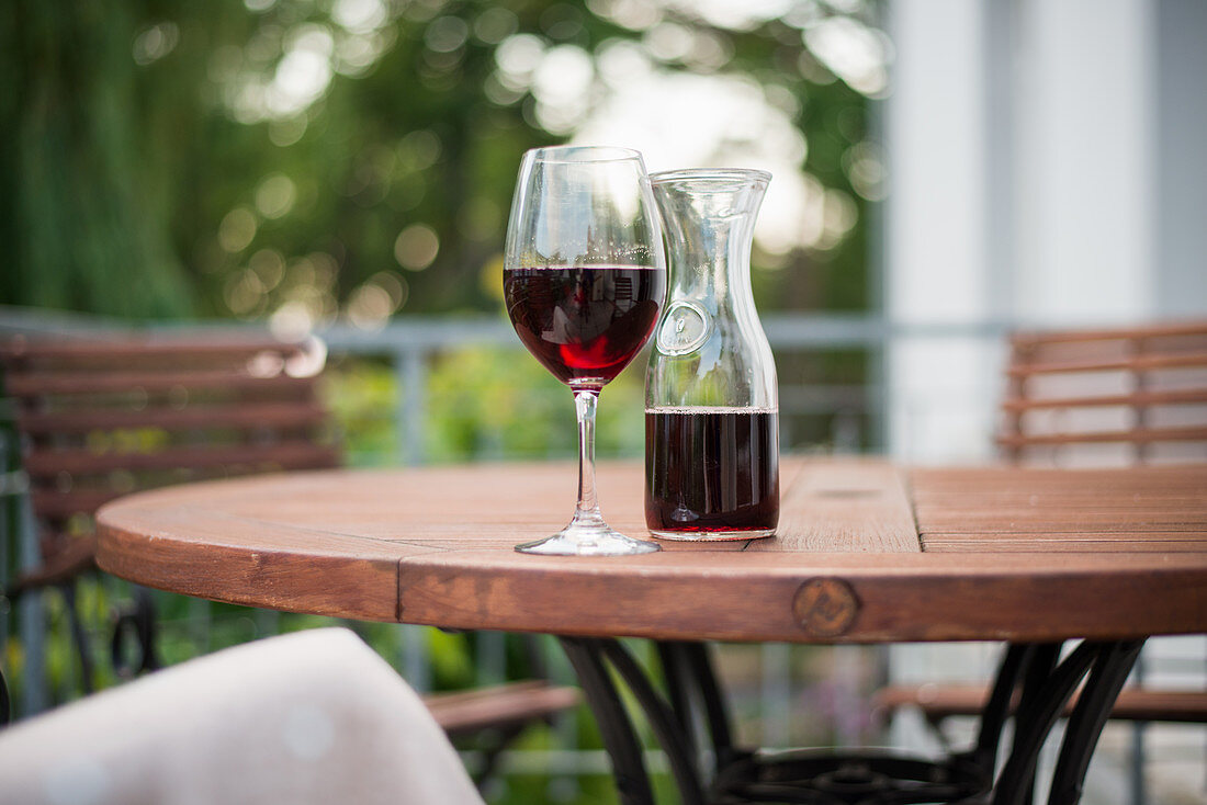 A glass of red wine and a carafe of wine on a wooden table in a garden