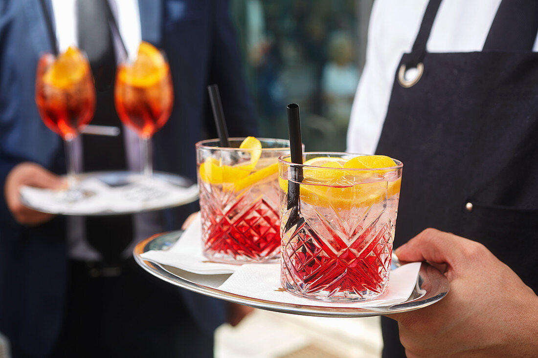 A waiter carrying cocktails on a tray (Campari Soda and Aperol Spritz)