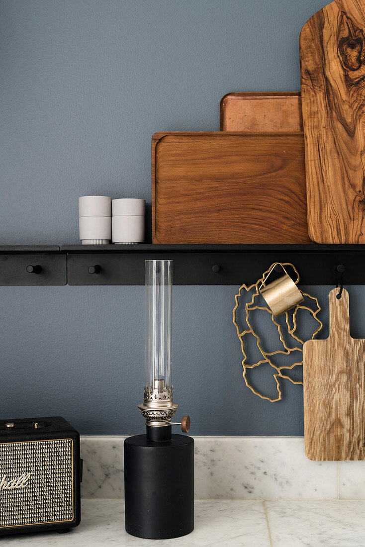 Wooden boards and trays on black ledge, oil lamp on marble worksurface and blue-grey wall in kitchen