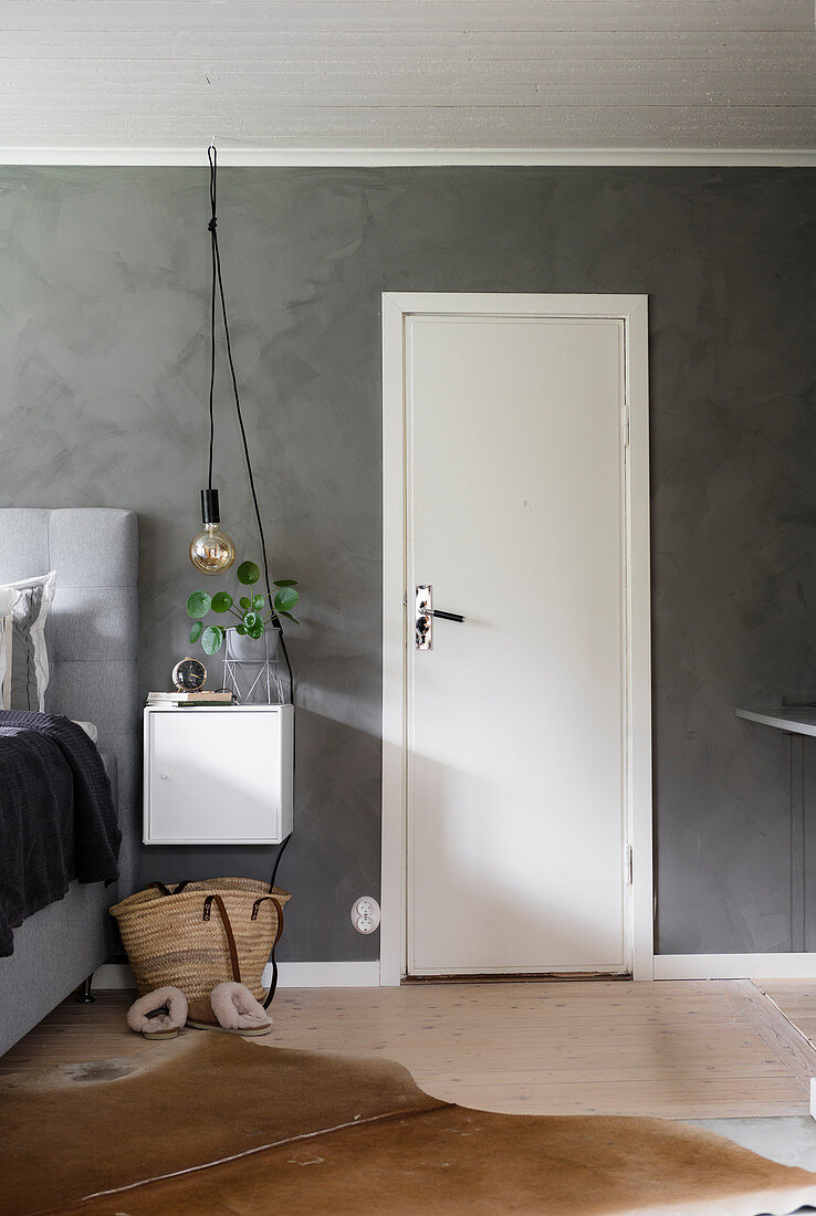 Pendelleuchte über Nachtkästchen, daneben weiße Tür im Schlafzimmer mit grauer Wand