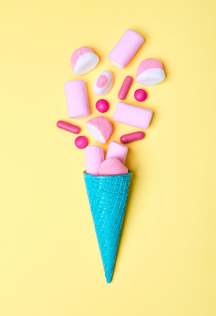 Bright blue ice-cream waffle cone with heap of pink candies and marshmallow on yellow background