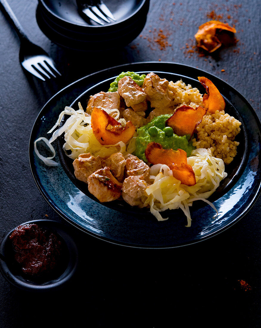 A bowl with Tandoori chicken, mushy peas, white cabbage and sweet potato crisps