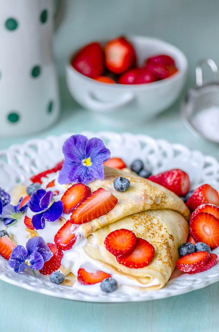 Thin pancakes with cream, strawberries and blueberries, decorated with violets for breakfast