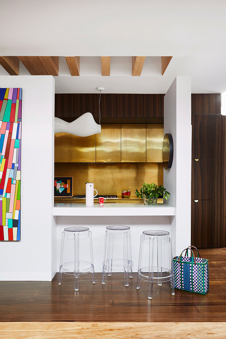 Kitchen with gold-colored fronts and back wall in front of breakfast counter with ghost stools