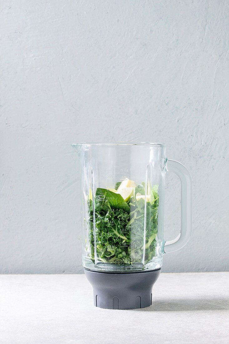 Ingredients for green spinach kale apple smoothie in glass blender on white marble table