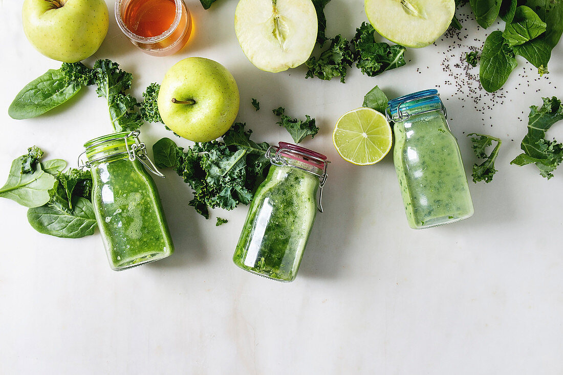 Variaty of green spinach kale apple honey smoothies in glass bottles with ingredients above over white marble background