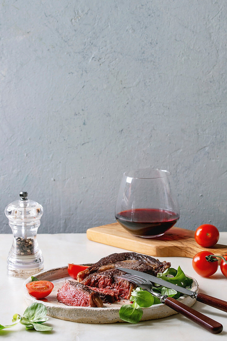 Grilled roasted sliced medium rare beef steak served in ceramic plate with green field salad, cherry tomatoes, cutlery, pepper, glass of red wine