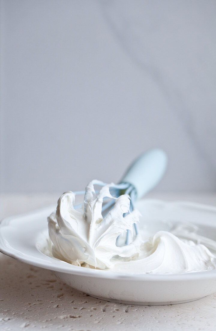 A blue whisk with icing, sitting in a white bowl on a white surface