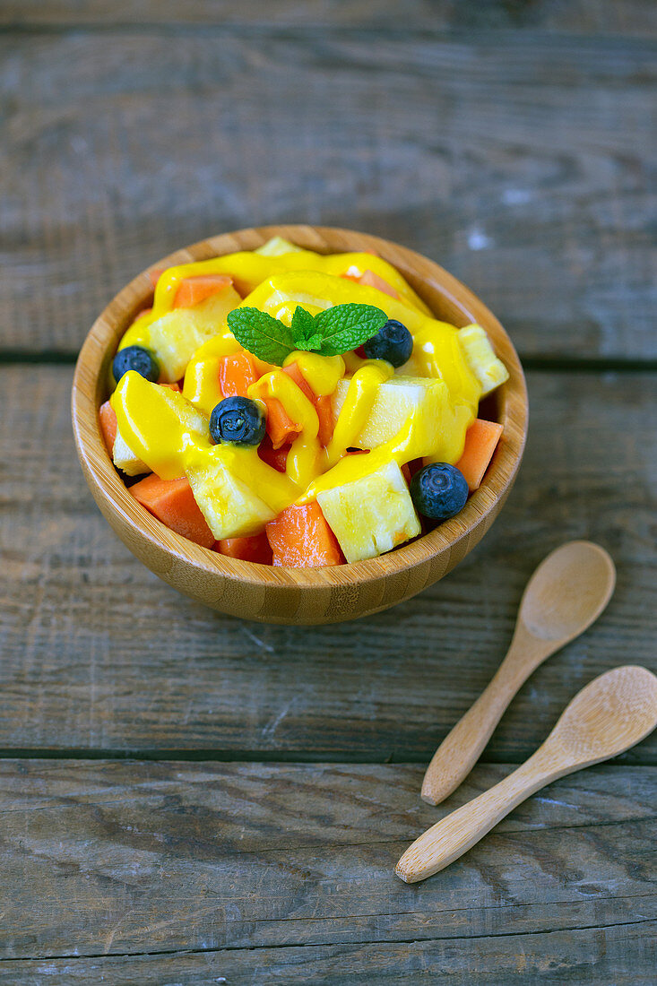 Juicy pieces of papaya with blueberries garnish by yellow sauce and mint leaf in bowl beside wooden spoons