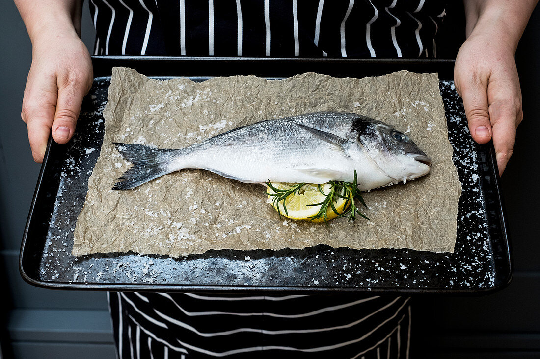 Person holding baking tray with fresh sea bream, slices of lemon and rosemary sprigs
