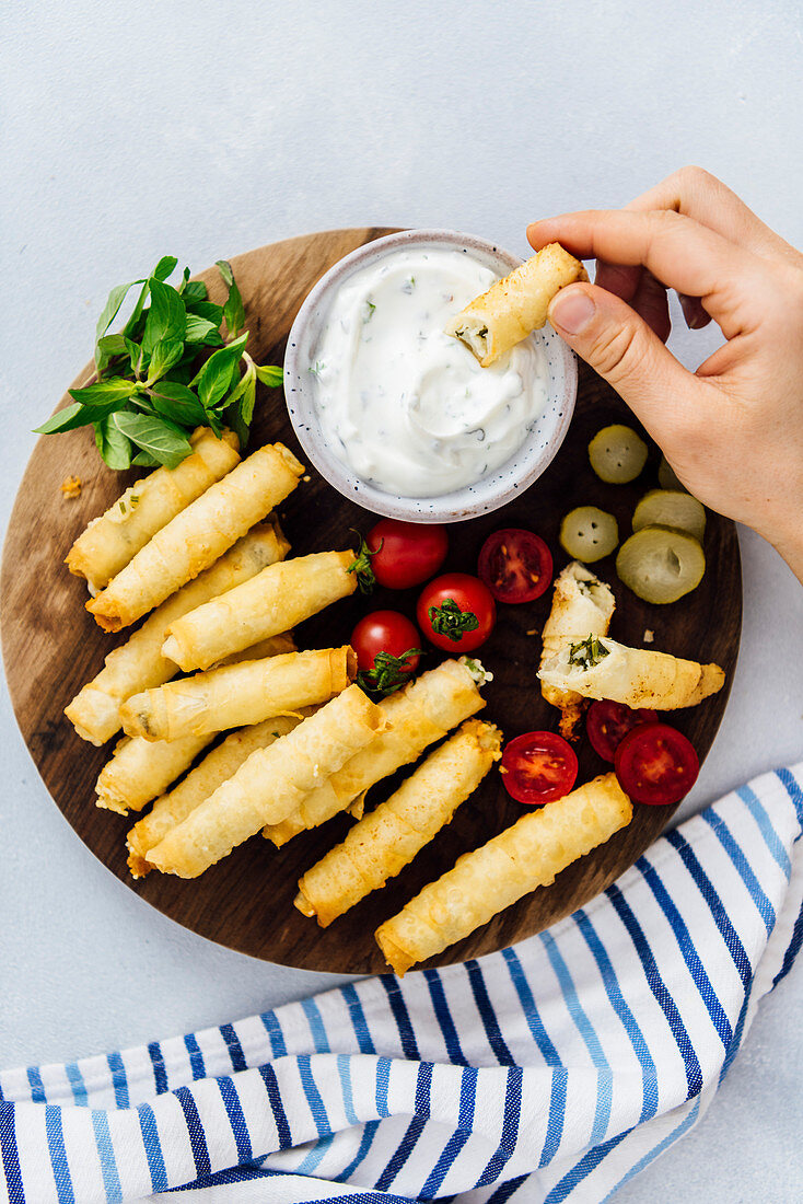 Woman eating stuffed phyllo rolls dipping into a yogurt sauce, tomatoes, mint and pickles accompany