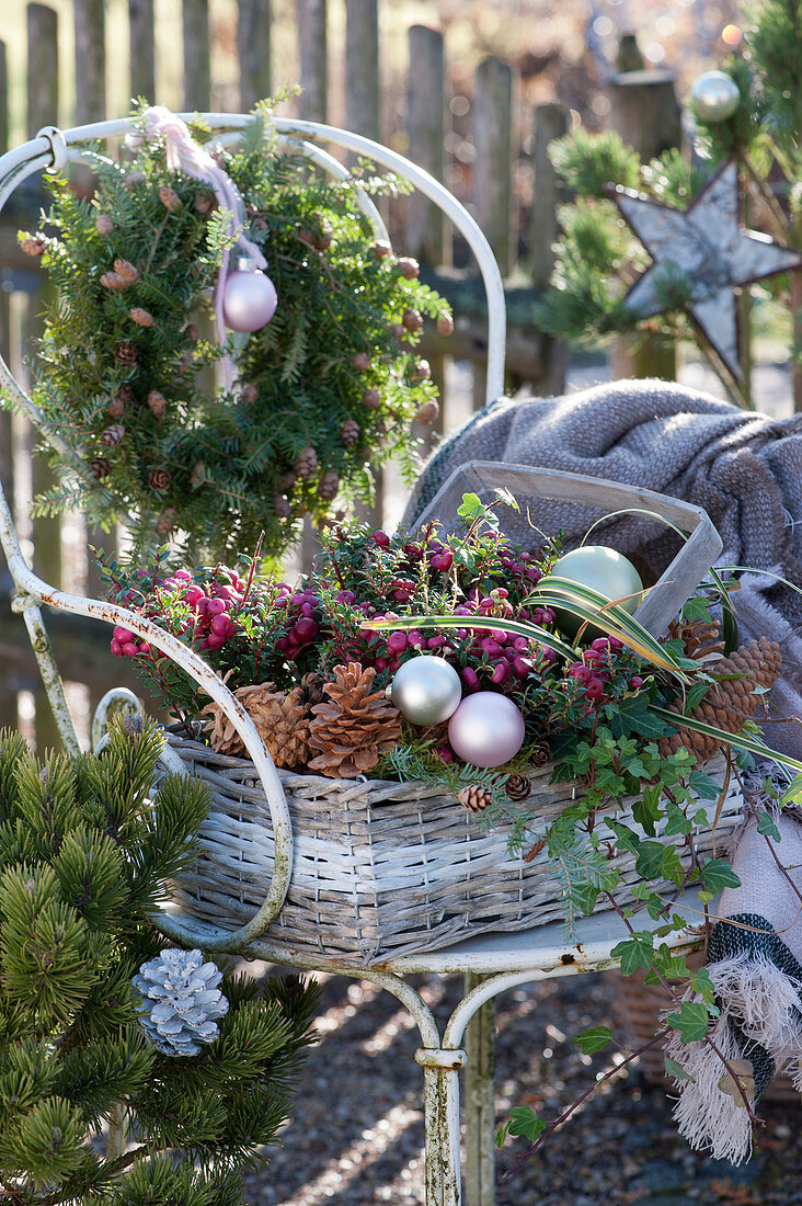 Basket with prickly heath, ivy, pinecones, and ornaments placed on a chair, hemlock wreath hanging on the back of the chair