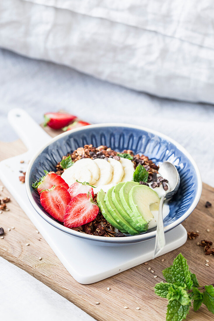 Almond and sesame seed granola with strawberries and avocado on a wooden board on a bed