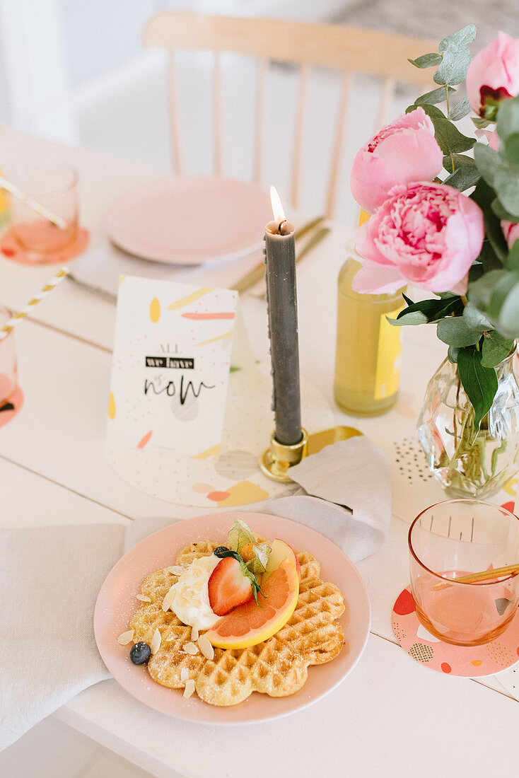 Waffel mit Früchten auf festlich gedecktem Tisch mit Kerze und Pfingstrosenstrauß