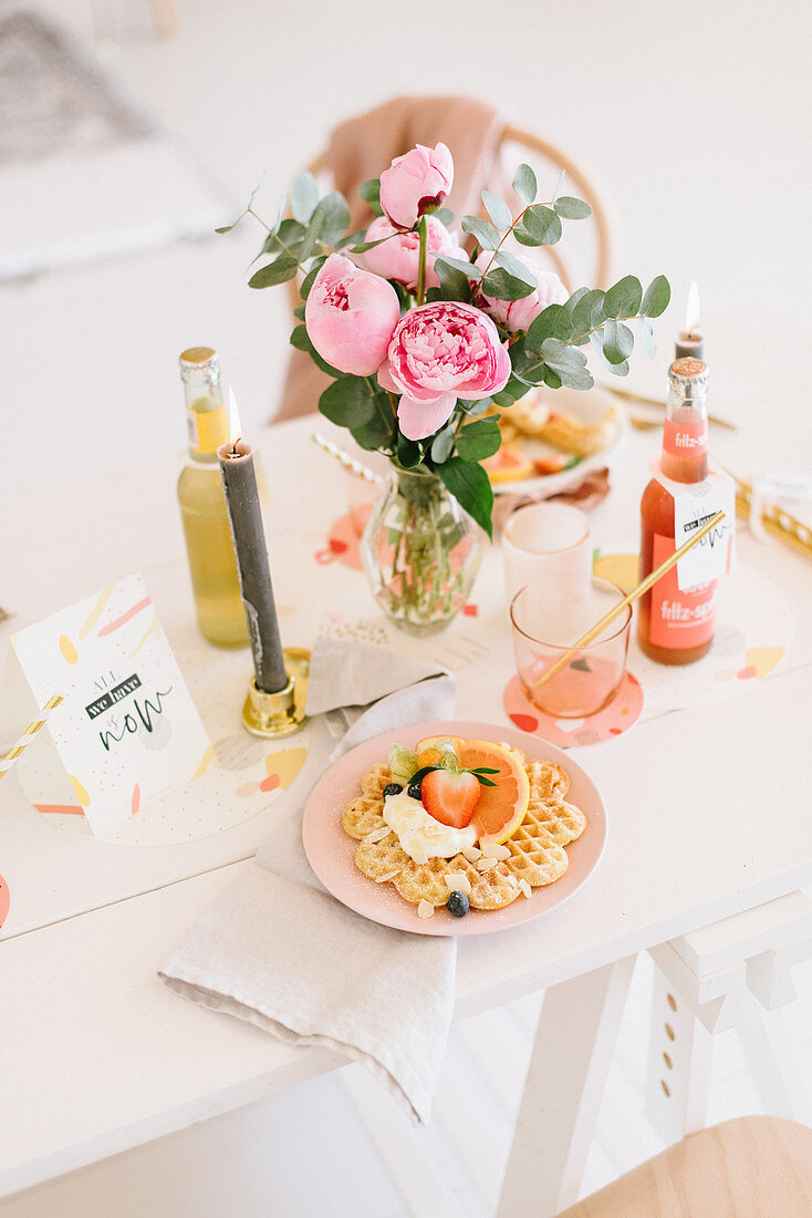 Waffles and fruit on table festively set with candle and vase of peonies