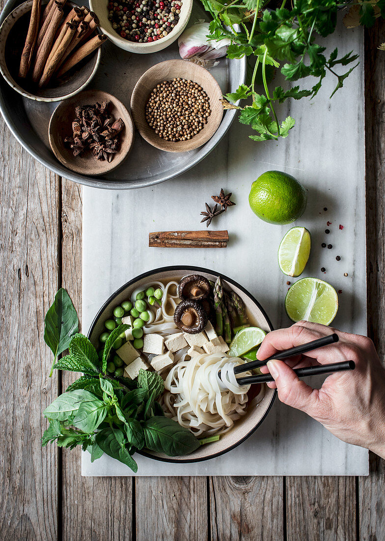 From above person hands with arranged bowls with dry spices and served Pho soup with noodles on marble board