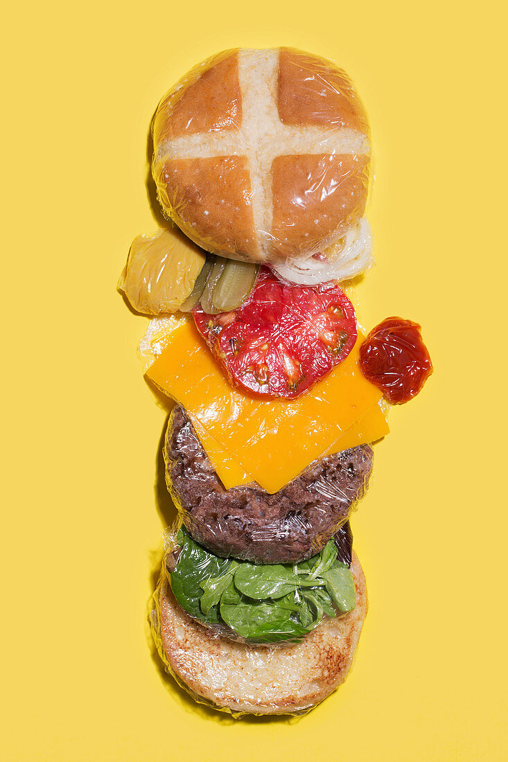 Different ingredients of a cheese burger wrapped in plastic on yellow background