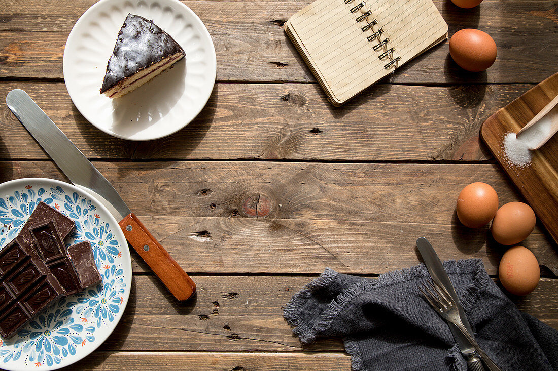 Stilleben mit Kuchenstück, Backzutaten und Notizblock auf rustikalem Holztisch