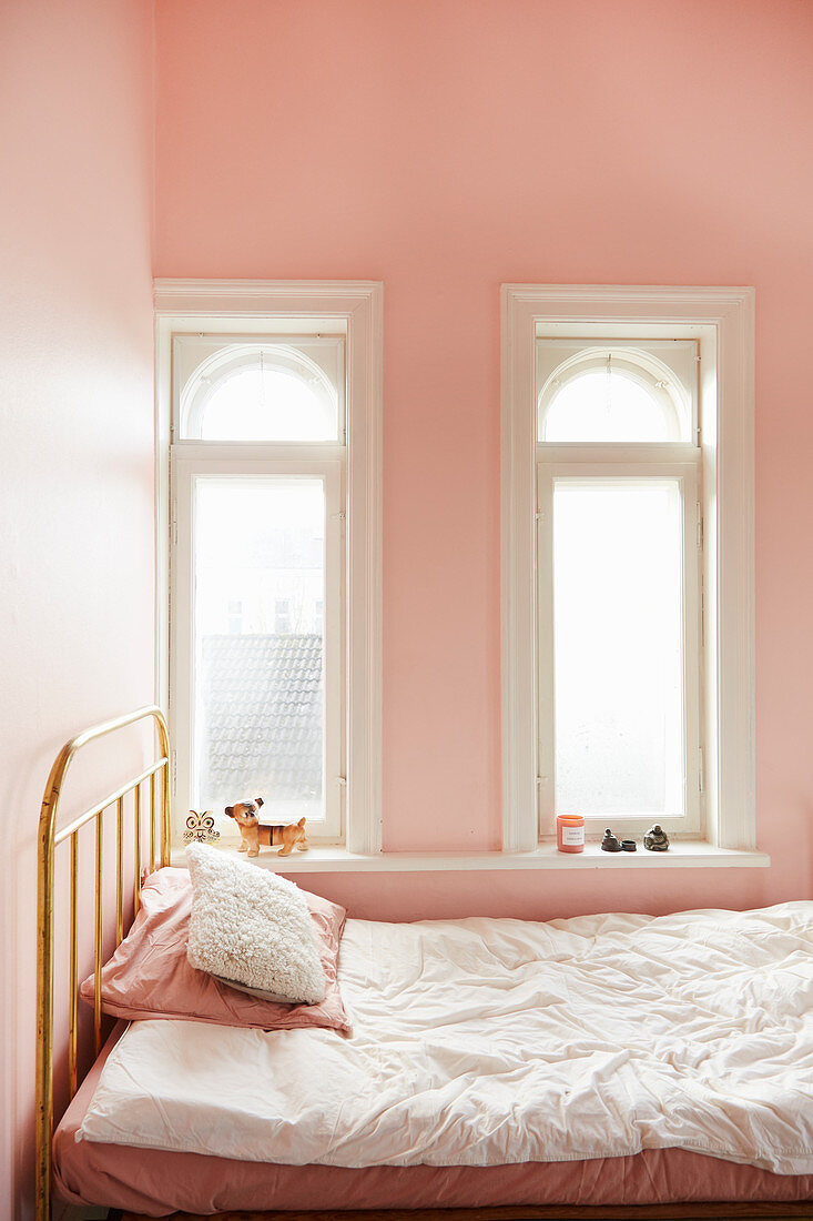 Metal bed below two small arched windows in pink bedroom
