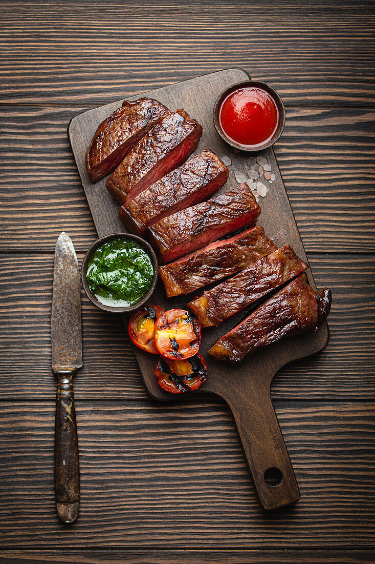 Grilled or fried and sliced marbled meat steak with fork, tomatoes as a side dish and different sauces on wooden cutting board, top view, close-up