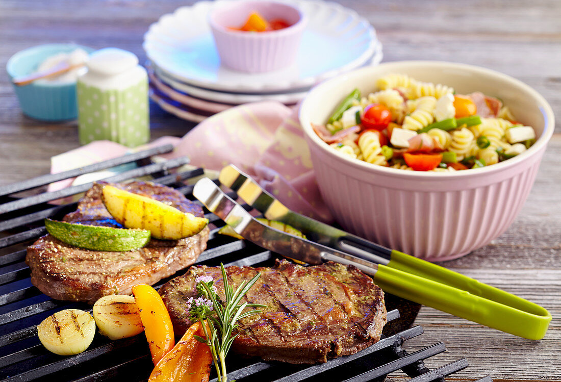 Marinated beef steaks with vegetables on a grill rack and a pasta salad