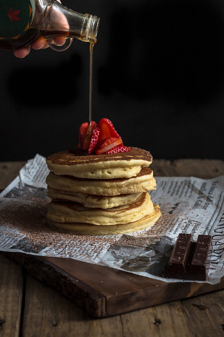 Person pouring maple syrup on appetizing tasty stack of pancakes with ripe strawberry