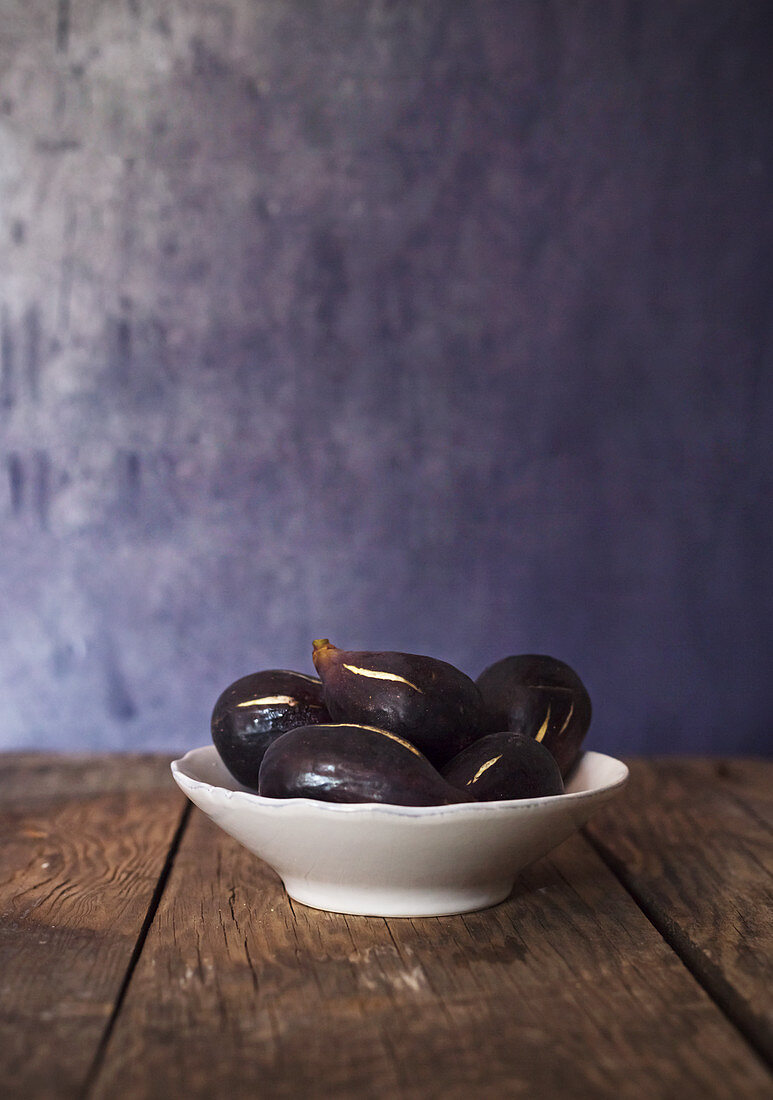 Ceramic bowl of ripe healthy figs placed on aged wooden table against blue wall