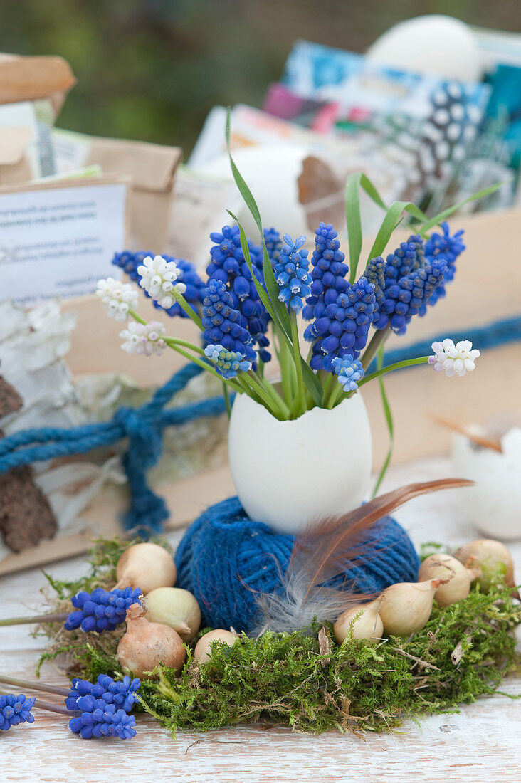 A small bouquet of grape hyacinths in a goose egg as a vase, spool of thread in a moss wreath with onions and feather