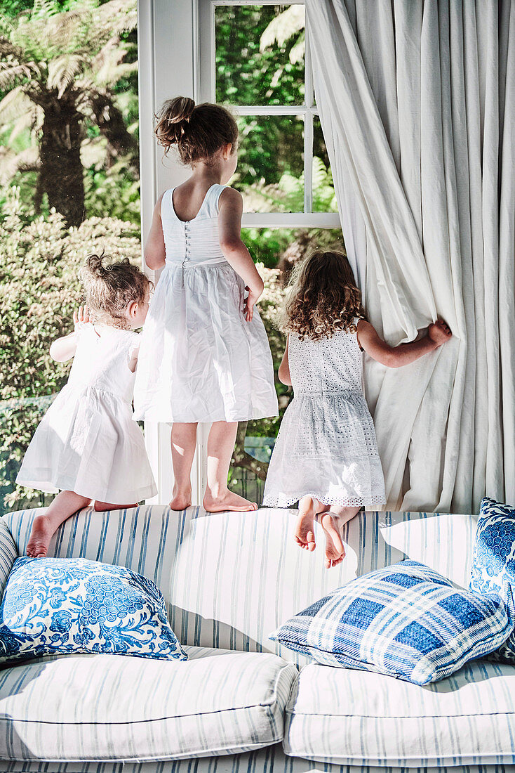 Drei Mädchen in weißen Sommerkleidern klettern auf Sofa am Fenster herum