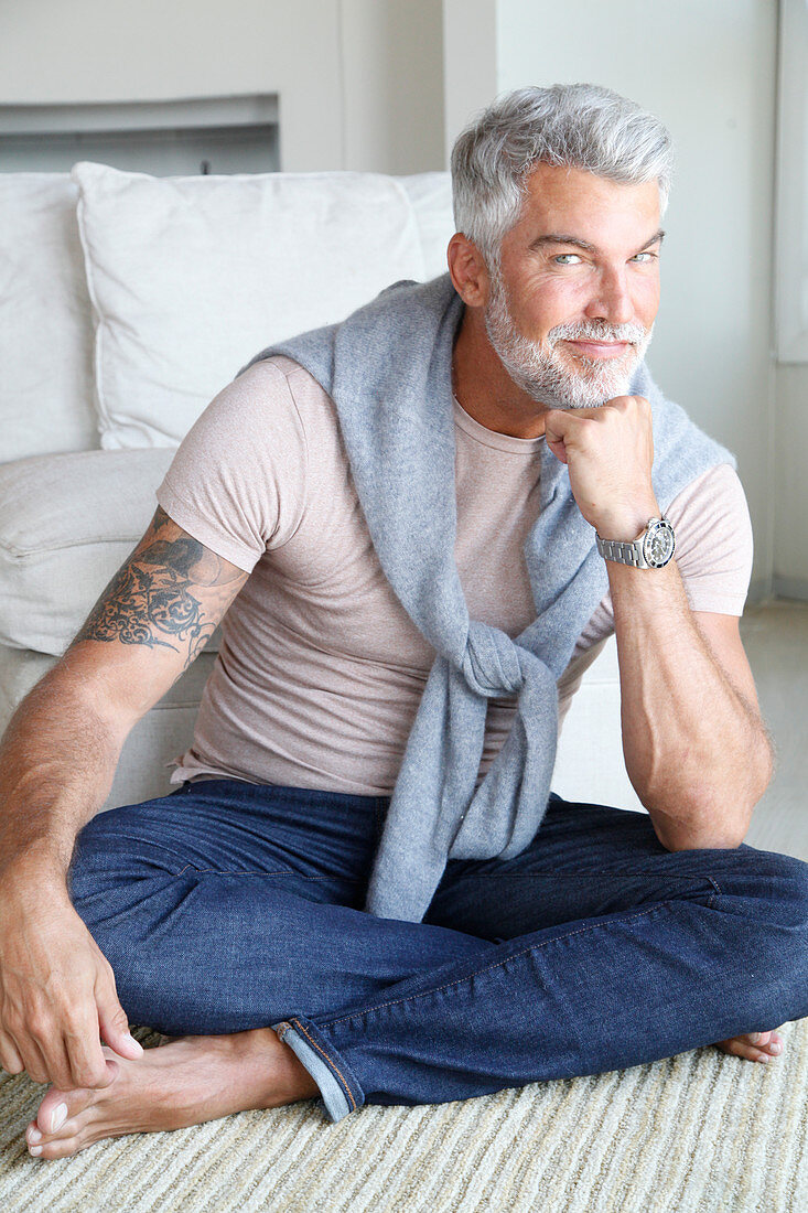 A grey-haired man wearing comfortable clothing sitting cross-legged on a bed