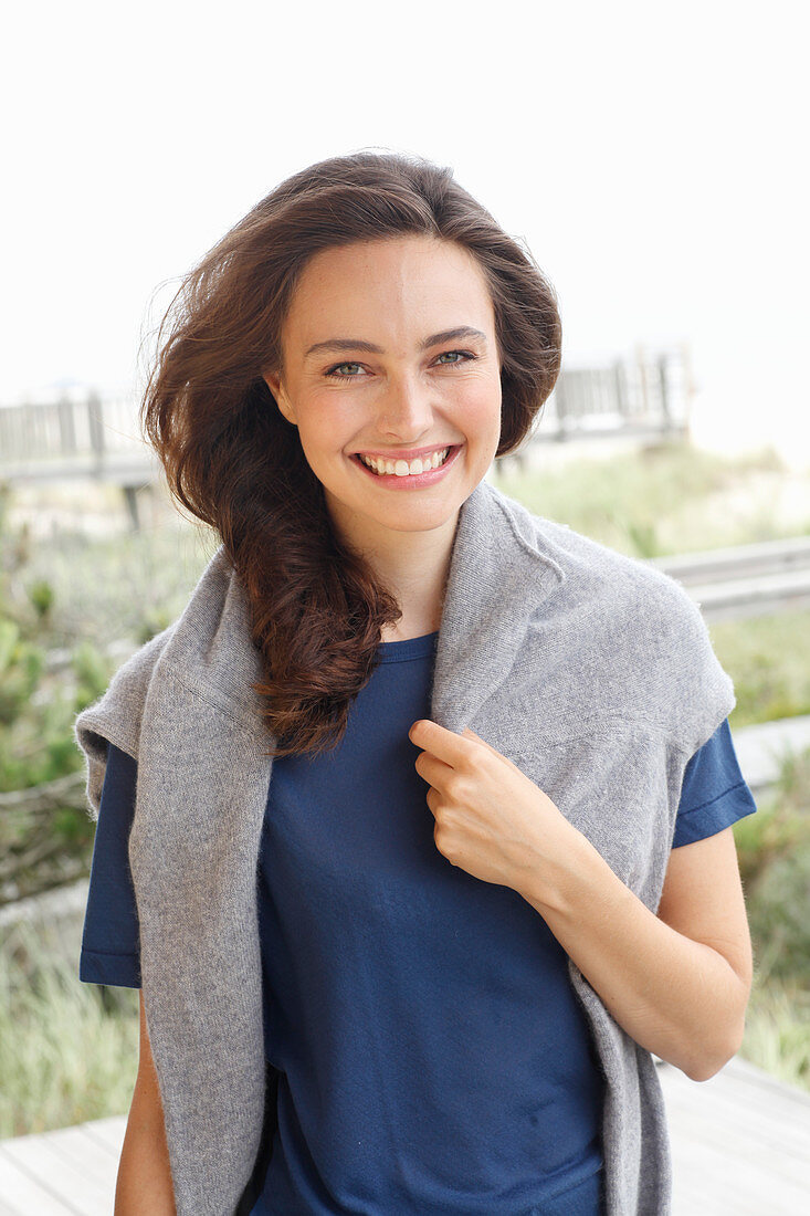 A young brunette woman wearing a short-sleeved blue t-shirt with a grey jumper around her shoulders