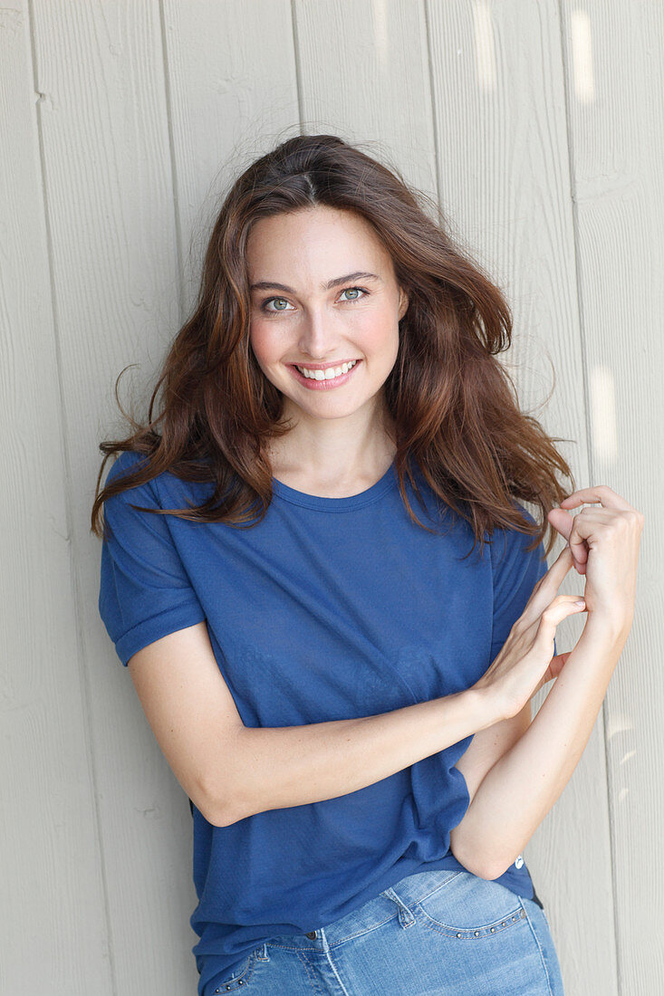 A young brunette woman in front of a grey wooden wall wearing a blue t-shirt