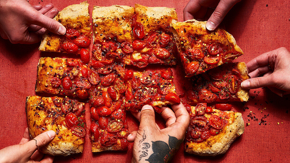 Roman style pizza topped with tomatoes and crushed red pepper flakes with hands (one tattooed)