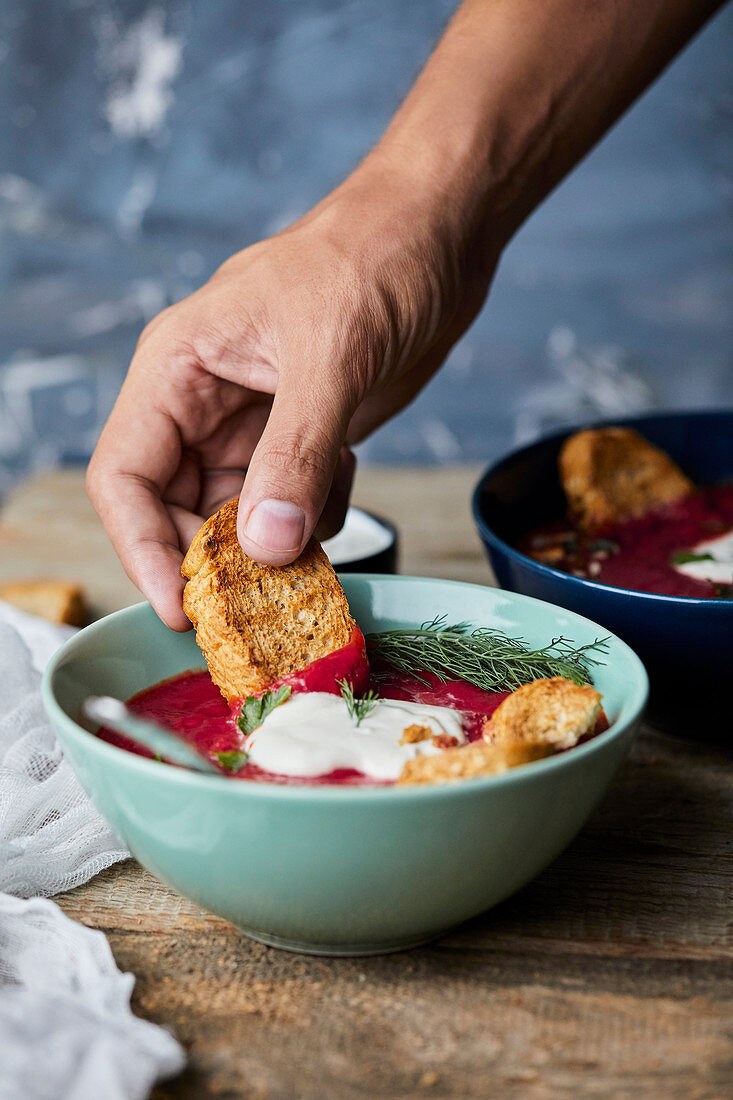 Hand dipping bread in bowl of Beetroot soup with coconut milk and sour cream