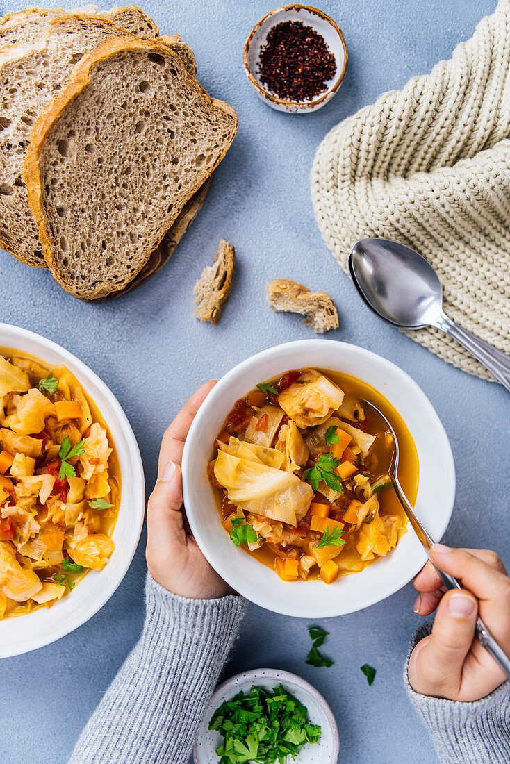 Woman having vegan cabbage soup in a white bowl, another bowl of soup, bread slices, chili flakes in a small ceramic bowl, parsley and spoons on the side photographed with a comforting winter feeling.