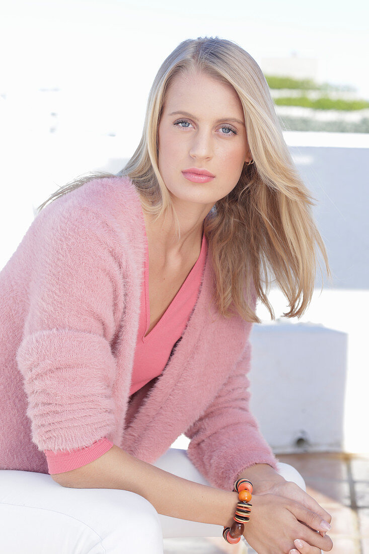 A young blonde woman wearing a pink top, a wrap-around jumper and white trousers