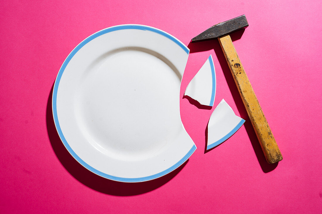 Broken white plate on pink background with hammer